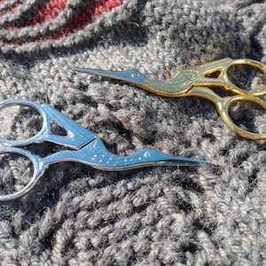 May include: Two pairs of decorative scissors, one silver and one gold, are lying on a gray knitted fabric. The silver scissors have a bird design on the handle.