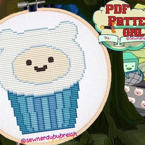 Peut inclure: Un motif de point de croix de Finn l'humain d'Adventure Time, dans un costume de cupcake bleu et blanc. Le motif est dans un cercle de broderie en bois. Le texte "PDF PATTERN ONLY" est dans une bulle de dialogue au-dessus du cercle. Le texte "by: @sewnerdybybreigh" est sous la bulle de dialogue.