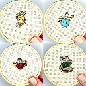 May include: Four embroidered hoops with different potion bottle pins. The pins are yellow, blue, red, and green. The pins have text on them that reads "Felix Felicis", "Veritaserum", "Amortentia", and "Polyjuice Potion".
