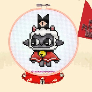 May include: Cross stitch pattern of a black and white sheep wearing a red cape and a gold bell. The pattern is in a hoop and the text "@sewnerdybybreich" is below the hoop.