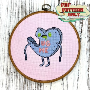 Op de afbeelding: Een borduurring met kruissteek met een roze achtergrond en een blauw buitenaards wezen met de tekst "Hug Me" op zijn borst.
