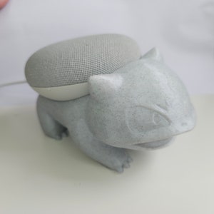 Può includere: Un personaggio Pokemon di colore grigio, testurizzato, stampato in 3D, che ricorda un Snorlax, tiene un altoparlante intelligente Google Home Mini grigio.