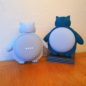 Può includere: Due dispositivi altoparlanti a forma di personaggio dei cartoni animati, uno grigio chiaro e uno verde acqua. Gli altoparlanti hanno un frontale rotondo in rete e sono su una superficie di legno. L'altoparlante grigio chiaro ha quattro luci bianche accese.