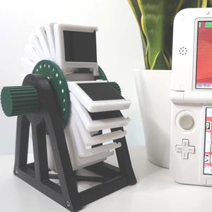 May include: A 3D-printed slide viewer in black, white, and green. The viewer features a rotating wheel design with multiple white slide holders. A white handheld gaming device is in the background.