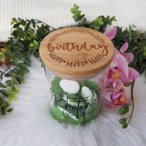 Könnte beinhalten: Ein Glasgefäß mit Bambusdeckel gefüllt mit grünen und weißen Süßigkeiten. Der Deckel hat die Worte "Happy Birthday" eingraviert. Das Gefäß hat auch die Worte "ein paar Kröten für dich" aufgedruckt.