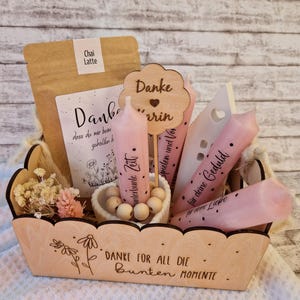 Könnte beinhalten: Ein hölzerner Geschenkkorb gefüllt mit verschiedenen Artikeln. Er enthält eine Tüte Chai Latte, eine Karte, rosa Kerzen mit Text, Holzdekorationen und Trockenblumen. Der Korb hat einen gewellten Rand und ein Blumendesign.