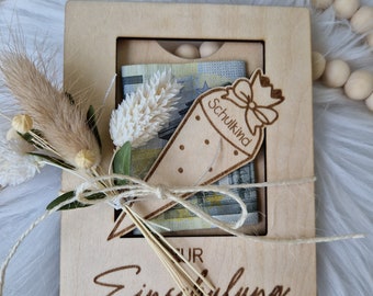 May include: A wooden box with a cutout for a school-themed gift. The box is decorated with the words "Zur Einschulung" and a small wooden schoolchild figure. The box contains a gift of money.