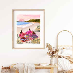 May include: A framed watercolor print depicts a beach picnic scene. Three figures in patterned clothing sit on a red blanket, enjoying food and drink. A wicker basket and a bottle of wine are visible. The background shows a beach, ocean, and sunset.