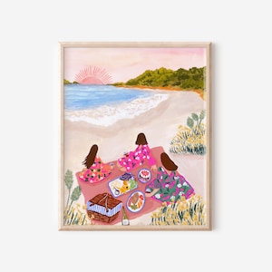May include: A framed art print depicts a beach picnic scene. Three figures in floral dresses sit on a pink blanket, enjoying food and drinks. A picnic basket, food platters, and a bottle of wine are visible. The background shows a beach, ocean, and sunset.