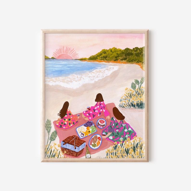 May include: A framed art print depicts a beach picnic scene. Three figures in floral dresses sit on a pink blanket, enjoying food and drinks. A picnic basket, food platters, and a bottle of wine are visible. The background shows a beach, ocean, and sunset.