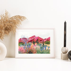 Op de afbeelding: Een ingelijste print van een woestijnlandschap met roze bergen, cactussen en kleurrijke bloemen. De print is in een wit frame.