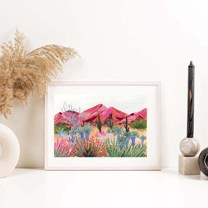 Op de afbeelding: Een ingelijste print van een woestijnlandschap met roze bergen, cactussen en kleurrijke bloemen. De print is in een wit frame.