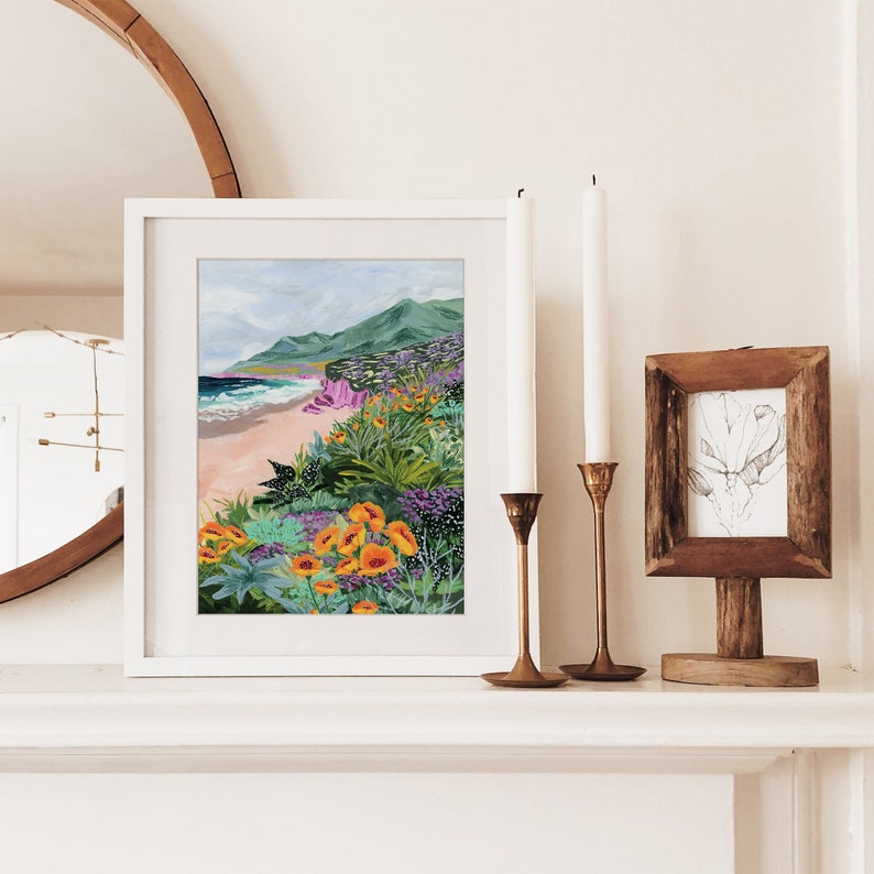 K&ouml;nnte beinhalten: Ein gerahmter Kunstdruck eines K&uuml;stenlandschaftsbildes mit Strand, Meer und Bergen. Das Kunstwerk zeigt leuchtende Farben, darunter Orange, Lila und Gr&uuml;n, die Wildblumen und Bl&auml;tter darstellen. Der Druck befindet sich auf einem wei&szlig;en Kaminsims mit Kerzen und einem kleinen botanischen Druck.