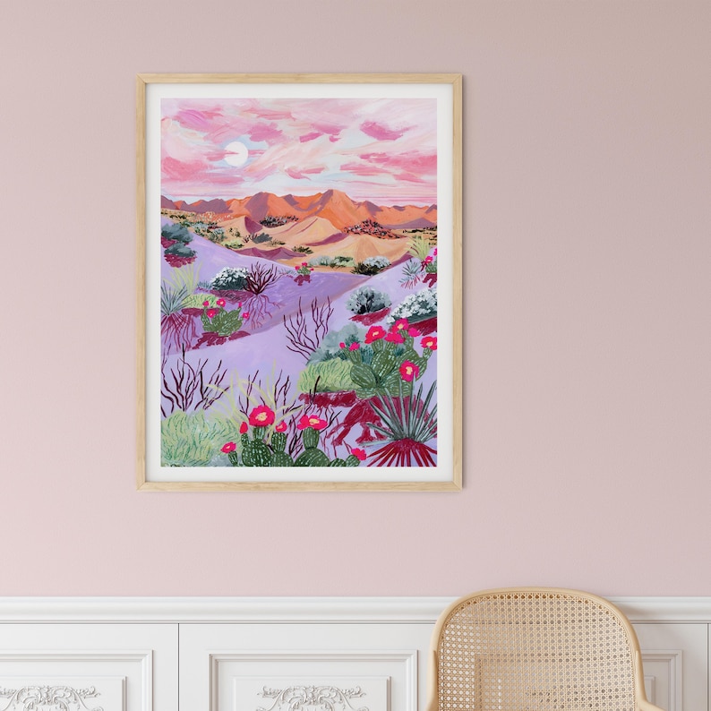 May include: A framed art print shows a desert scene with a pink and purple sky, a full moon, and orange mountains. The foreground features cacti with red flowers and various desert plants in shades of green and purple.