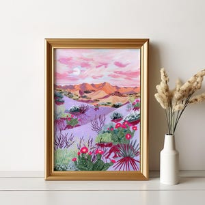 May include: A framed desert landscape painting in a gold frame. The artwork shows a desert scene with pink and orange mountains, a purple foreground with cacti and flowering plants, and a pink sky with a moon.