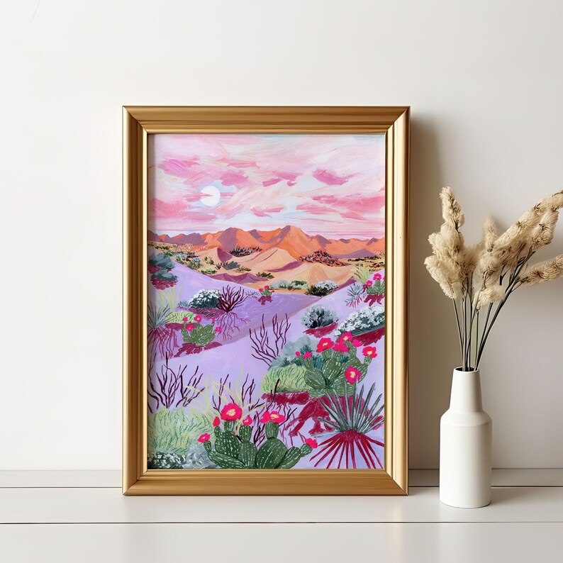 May include: A framed desert landscape painting in a gold frame. The artwork shows a desert scene with pink and orange mountains, a purple foreground with cacti and flowering plants, and a pink sky with a moon.