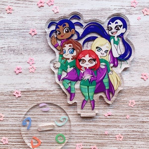 May include: Five cartoon characters in a group, standing on a clear acrylic charm. The characters are all wearing green and purple clothing and have different hair colors. The charm is surrounded by pink glitter stars.