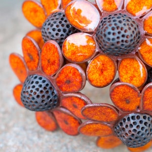 Könnte beinhalten: Ein handgefertigter Blumenring mit orangefarbenen und schwarzen Blütenblättern. Der Ring hat ein einzigartiges Design mit einem Cluster von Blumen, jede mit einem strukturierten, perforierten Zentrum. Die Blütenblätter sind leuchtend orange.