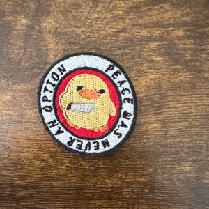 Peut inclure: Un écusson rond brodé avec un fond blanc et un contour noir. Le centre de l'écusson présente un cercle rouge avec un poulet jaune de dessin animé tenant un couteau. Le texte "NEVER" est écrit en noir sur le cercle rouge. Le texte "WAS" est écrit en noir sur le fond blanc. Le texte "PEACE" est écrit en noir sur le fond blanc. Le texte "AN OPTION" est écrit en noir sur le fond blanc.
