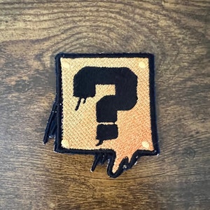 May include: A black and gold embroidered patch with a question mark inside a box. The box has a dripping effect.