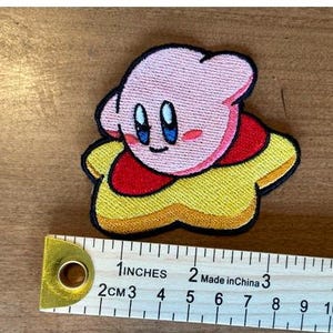 Peut inclure: Patch brodé rose et jaune représentant un personnage de dessin animé en forme d'étoile. Le personnage a de grands yeux bleus et un corps rose.