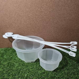 May include: Two clear, plastic, hexagonal-shaped containers of different sizes, along with several white plastic hanging plant supports. The containers are on a bed of green artificial grass, with a brown background.