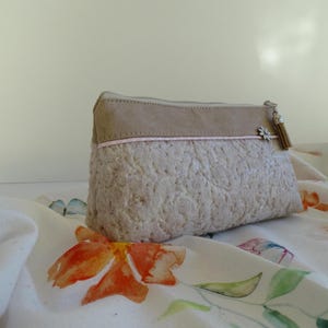 Op de afbeelding: Een beige en bruine toilettas met een kant-achtig patroon en een klein kwastje. De tas ligt op een witte stof met bloemenmotieven.