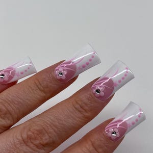 May include: A hand with long, square-shaped artificial nails. The nails are white with pink floral designs and small rhinestones. The design includes pink swirls and dots.