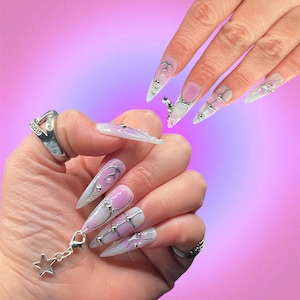 Peut inclure: Gros plan sur des mains avec de longs ongles en acrylique blancs et pointus d&eacute;cor&eacute;s de tourbillons argent&eacute;s et roses, de strass et d'un charme de cha&icirc;ne argent&eacute;e avec un pendentif en forme d'&eacute;toile.