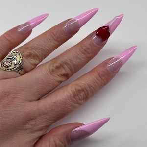 May include: A hand with long, pointed, pink acrylic nails. The nails have a glittery, light pink base and a darker pink tip. One nail features a red heart design. A gold ring with a floral design is on the ring finger.