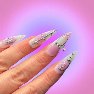 Peut inclure: Gros plan sur une main avec de longs ongles pointus, ombr&eacute;s de blanc et de rose, avec des paillettes argent&eacute;es et des strass. Les ongles sont d&eacute;cor&eacute;s d'un motif en spirale argent&eacute;.