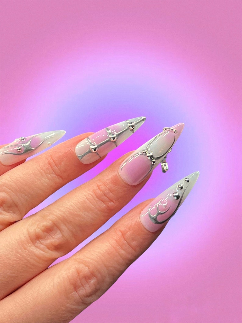 Peut inclure: Gros plan sur une main avec de longs ongles pointus, ombr&eacute;s de blanc et de rose, avec des paillettes argent&eacute;es et des strass. Les ongles sont d&eacute;cor&eacute;s d'un motif en spirale argent&eacute;.