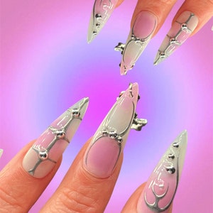 Peut inclure: Gros plan sur une main avec de longs ongles pointus. Les ongles sont peints avec un d&eacute;grad&eacute; de rose et de blanc, et d&eacute;cor&eacute;s de paillettes argent&eacute;es et de petites perles argent&eacute;es.