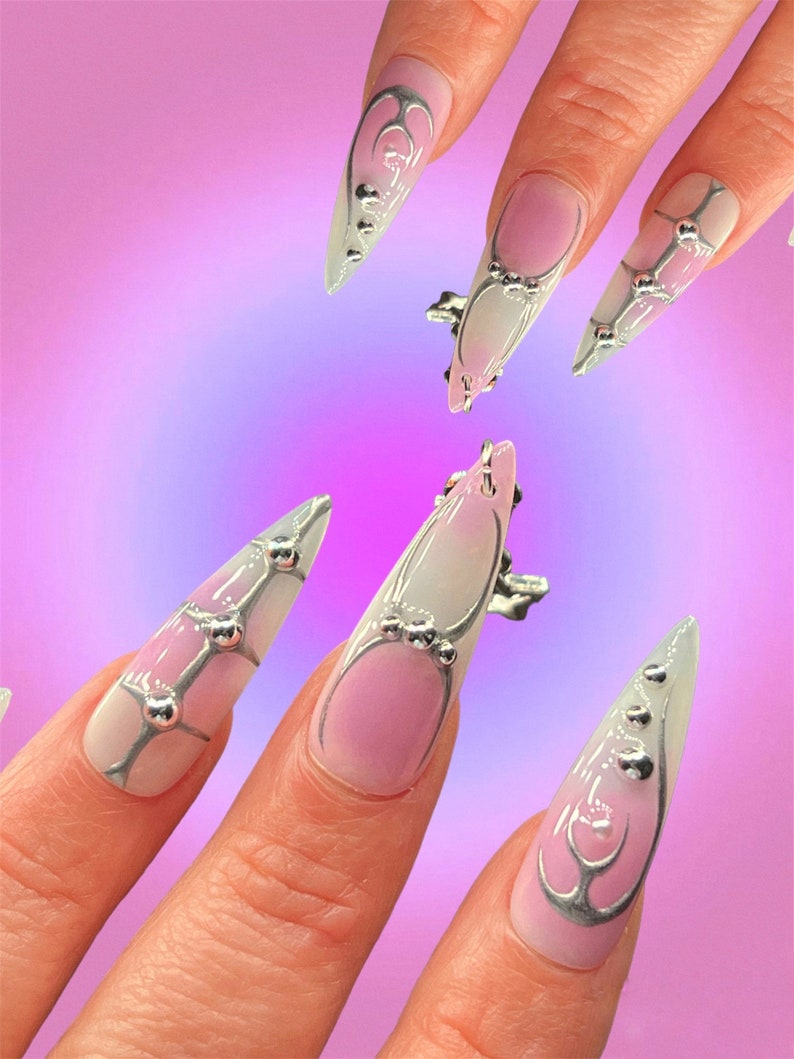 Peut inclure: Gros plan sur une main avec de longs ongles pointus. Les ongles sont peints avec un d&eacute;grad&eacute; de rose et de blanc, et d&eacute;cor&eacute;s de paillettes argent&eacute;es et de petites perles argent&eacute;es.