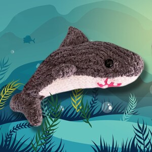May include: Two handmade crochet sharks featuring dark grey upper bodies and white underbellies. One shark is positioned horizontally in profile, while the other is angled vertically with its mouth open, revealing pink and white stitched teeth.