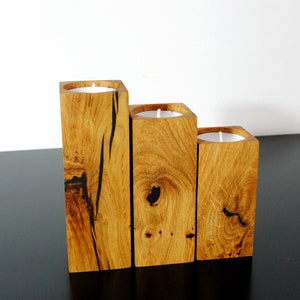 Könnte beinhalten: Drei Kerzenhalter aus Holz in abgestuften Größen, jeder mit einer schwarz gebeizten Oberfläche und einer runden Öffnung für ein Teelicht. Die Halter sind versetzt auf einer schwarzen Oberfläche angeordnet.