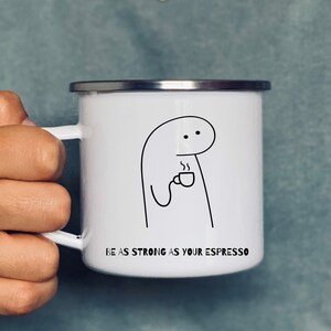 Op de afbeelding: Een witte emaille mok met een zwarte lijntekening van een persoon die een kopje koffie vasthoudt. De tekst "BE AS STRONG AS YOUR ESPRESSO" staat op de mok gedrukt.