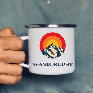 Può includere: Tazza da campeggio in smalto bianco con un grafico di tramonto rosso, arancione e giallo e una silhouette di montagna. La tazza ha un bordo e un manico neri e il testo "WANDERLUST" stampato sul davanti.