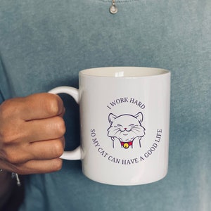 Könnte beinhalten: Weiße Keramiktasse mit einem schwarzen und violetten Design. Das Design zeigt eine Comic-Katze mit einem roten Halsband und einer Glocke und den Text "Ich arbeite hart, damit meine Katze ein gutes Leben haben kann".