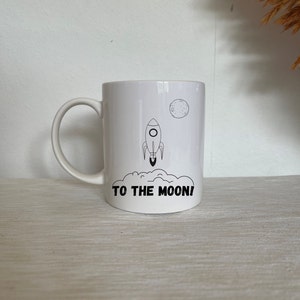 Może przedstawiać: Biały kubek ceramiczny z czarnym konturem rakiety startującej w kierunku czarnego księżyca. Tekst "TO THE MOON!" jest nadrukowany czarnym kolorem pod rakietą.