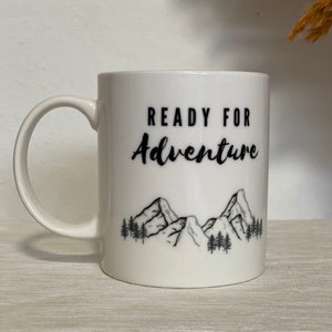 Może przedstawiać: Biały kubek ceramiczny z czarnym nadrukiem gór i drzew. Na kubku widnieje napis "Ready for Adventure".