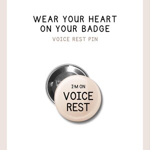 May include: A round pin-back button with a pale pink surface featuring the black text 'I'M ON VOICE REST'. The text 'WEAR YOUR HEART ON YOUR BADGE' and 'VOICE REST PIN' appear above the button. The pin has a metallic silver back visible behind it.