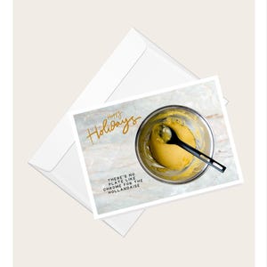 Op de afbeelding: Een kerstkaart met een gouden schrijflettertype in cursief dat "Prettige feestdagen" zegt. De kaart toont een foto van een kom met gele hollandaisesaus met een zwarte lepel erin. De tekst onder de foto luidt "Er is geen bord zoals chroom voor de hollandaise."