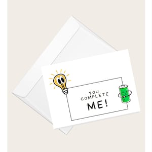 Può includere: Biglietto di auguri bianco con la scritta "YOU COMPLETE ME!" e un'illustrazione a cartoni animati di una lampadina e una batteria. Il biglietto è accompagnato da una busta bianca, perfetto per inviare un messaggio d'amore o apprezzamento.