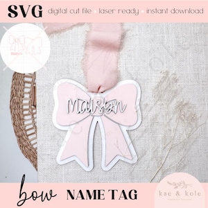 Può includere: Un'etichetta a forma di fiocco rosa con il nome "Maisan" in corsivo. L'etichetta ha un contorno bianco ed è appesa a un nastro rosa. Le parole "bow NAME TAG" sono in basso, con "SVG digital cut file laser ready instant download" in alto.