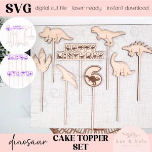 Op de afbeelding: Een set houten dinosaurus taarttoppers. De set bevat verschillende dinosaurusvormen, een "Happy Birthday" bord, cijfers en de tekst "Cake Topper Set". De toppers zijn ontworpen voor digitaal snijden en direct downloaden.