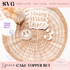 May include: A three-piece cake topper set with the words "Young Wild & Three", a smiley face, and the number "3". The toppers are made of wood and are perfect for a birthday party.