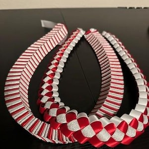 Può includere: Una collana di laurea realizzata con nastri piegati rossi, bianchi e argento glitterati. La collana presenta un complesso motivo intrecciato con anelli alternati di raso rosso tinta unita, raso bianco tinta unita e nastro argento glitterato.