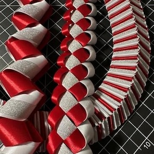 May include: Three decorative leis crafted from red satin ribbon, white satin ribbon, and silver glitter ribbon. The ribbons are folded and woven into intricate, layered patterns, creating a textured, multi-dimensional appearance with a festive look.