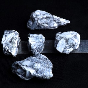 May include: Five grey and white rocks of varying sizes are displayed on a black background. A metal ruler is positioned alongside the rocks to show scale. The rocks have a rough, textured surface.